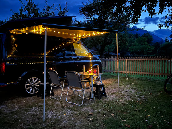 Camping van standing on a camp ground in the evening with a table and chairs infront of it and fairy lights glistening